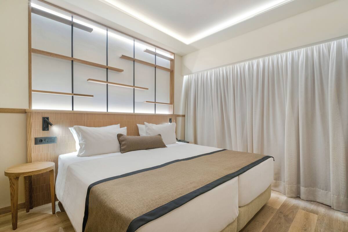 Ein Hotelzimmer mit einem großen Bett, einem Nachttisch, einem an der Wand montierten Fernseher und einem Fenster mit Vorhängen.