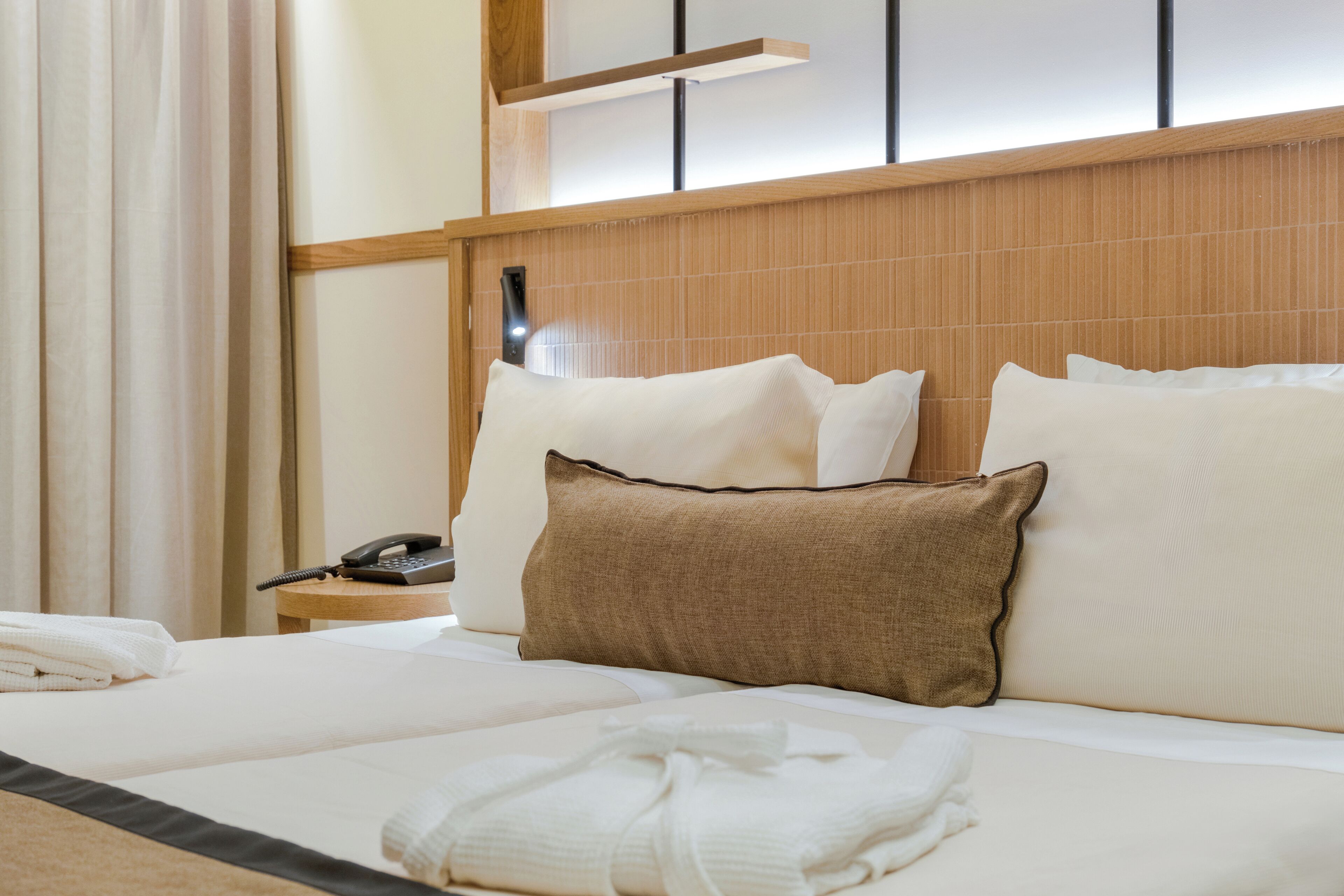 A neatly made bed with white linens and a brown pillow, a bedside telephone, and a wooden nightstand.