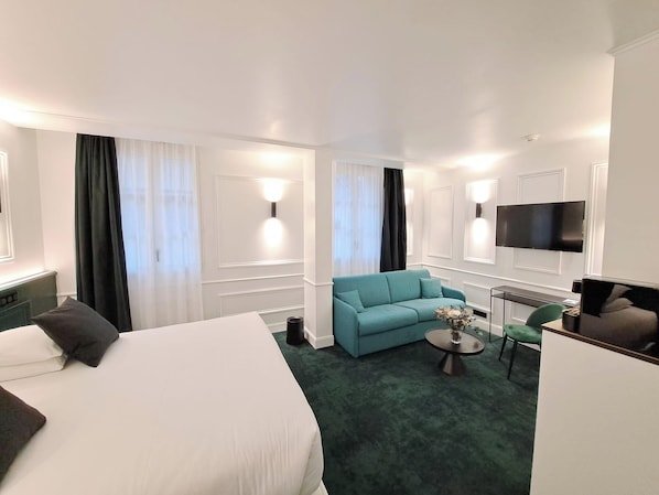 A hotel room with a large bed, a sofa, a small round table with a vase, a desk with a computer, and a TV mounted on the wall.