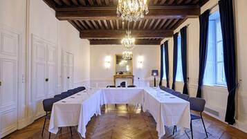 A conference room with a long table, chairs, a chandelier, wooden ceiling beams, and large windows with curtains.