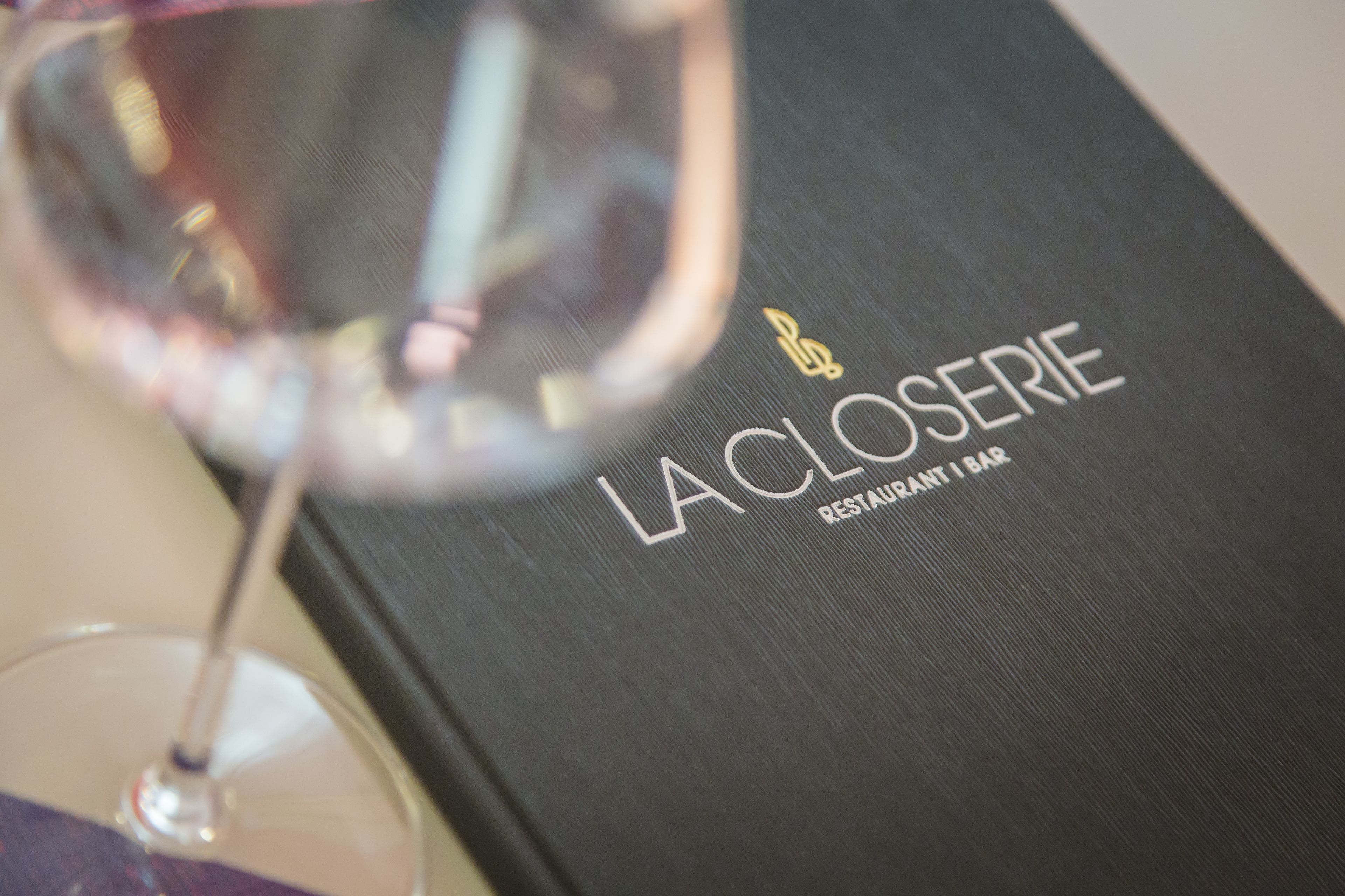 Gros plan sur un verre à vin et un livre noir portant l’inscription « LA CLOSERIE RESTAURANT BAR ».