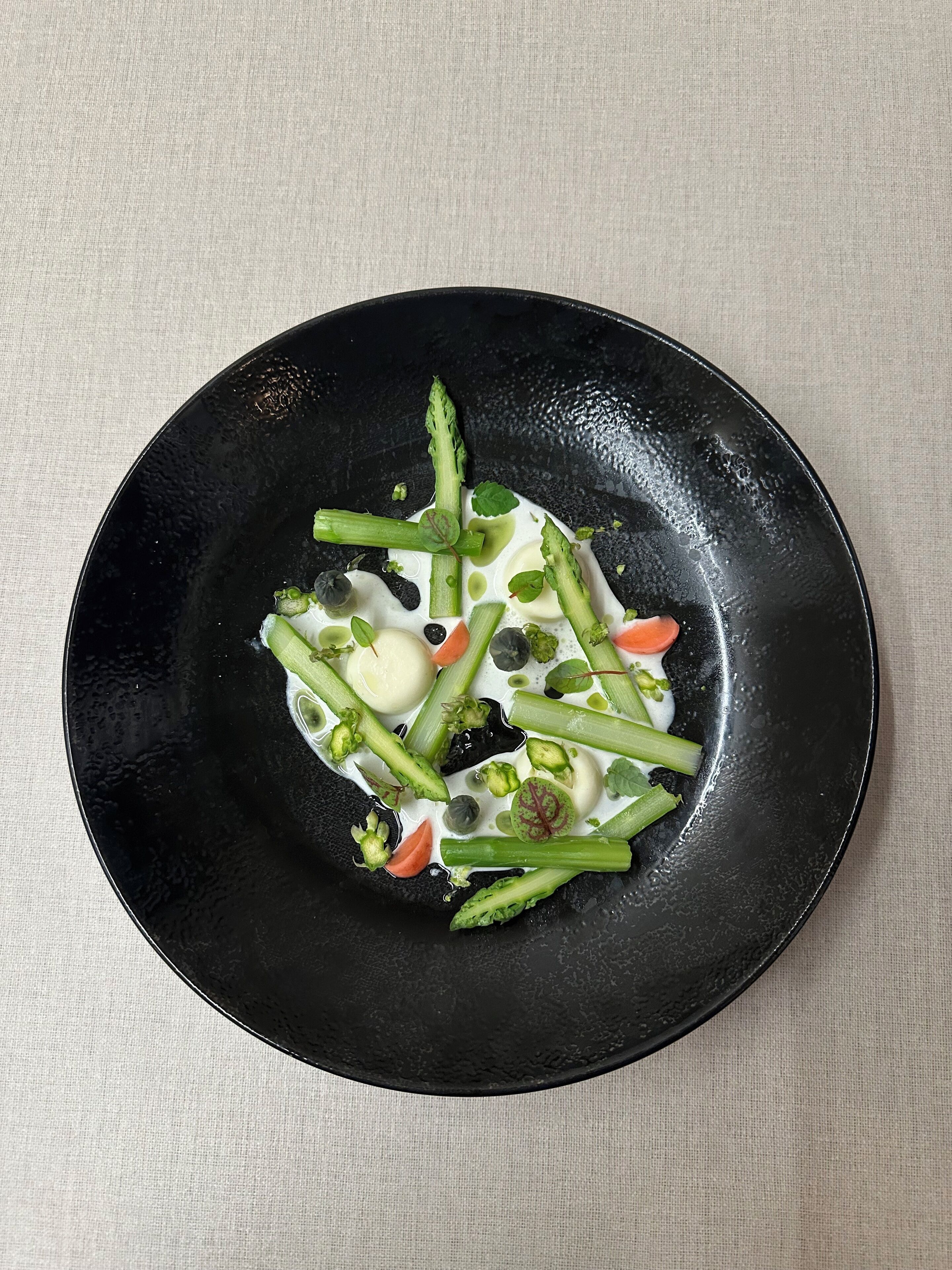 Un plato negro con salsa blanca, verduras verdes y una cucharada de crema sobre una superficie texturizada.