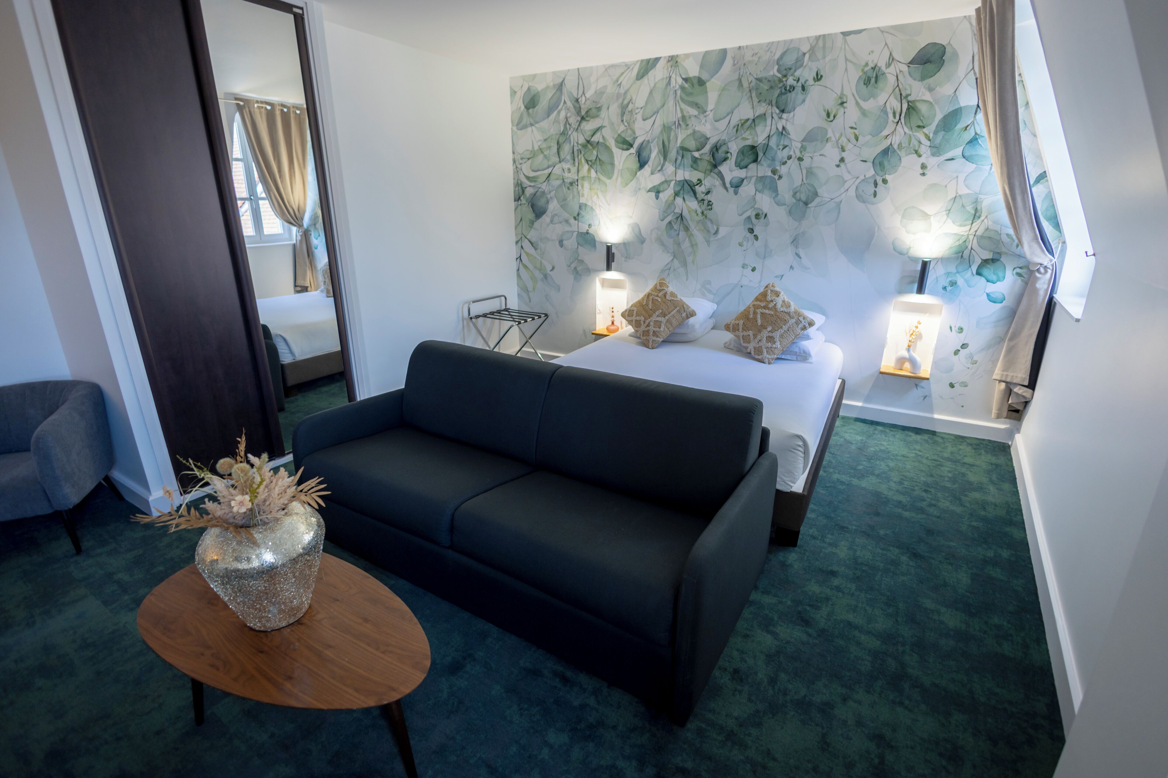 A hotel room with a bed, a dark-colored sofa, a small round table with a decorative vase, and a wall with a leafy pattern.