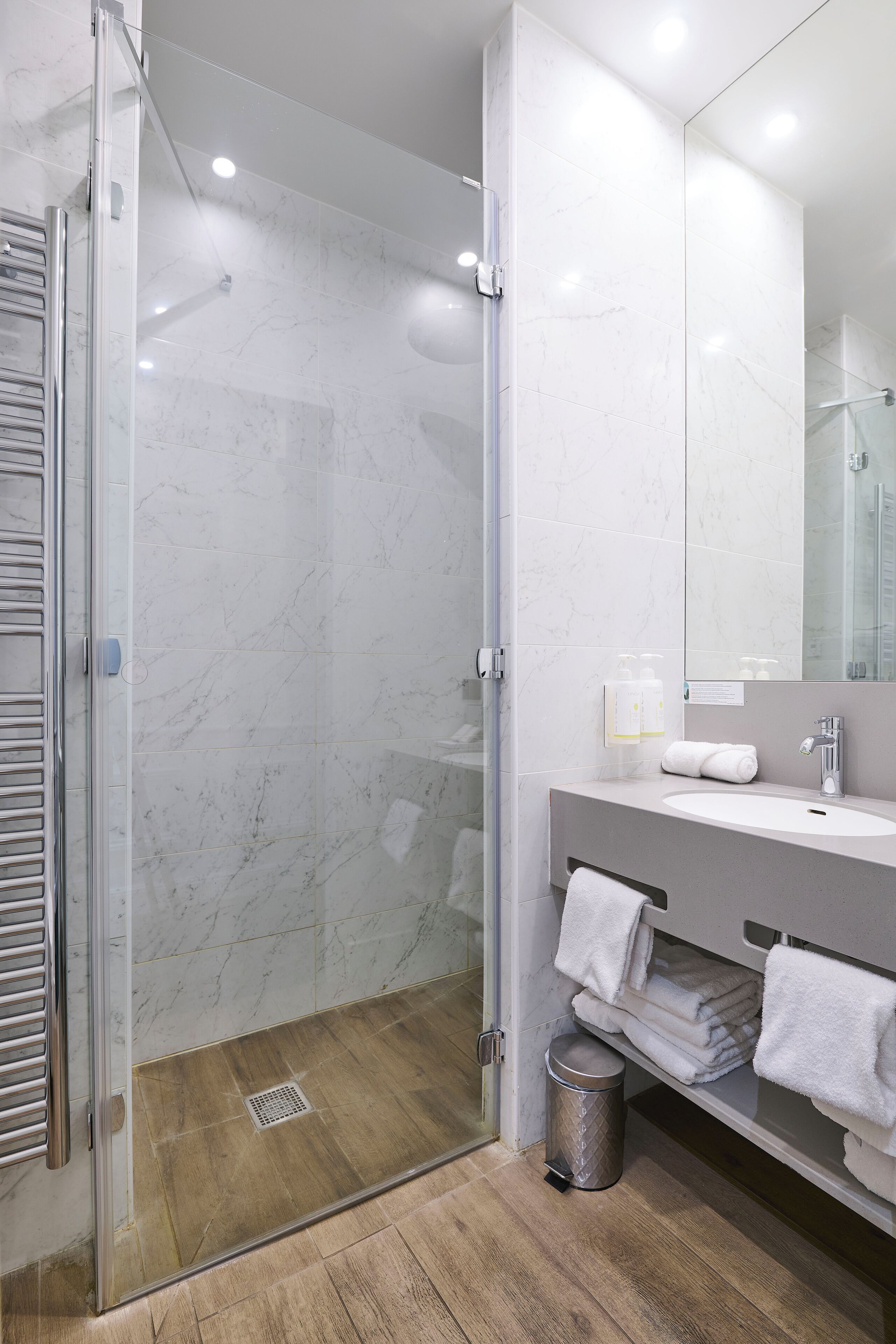 A modern bathroom with a glass-enclosed shower, a marble-tiled wall, a wooden floor, and a vanity with towels.