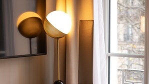 Habitación de hotel con un escritorio de madera, una silla verde azulada, una lámpara, una taza, un plato con un pastelito y una ventana con cortinas.