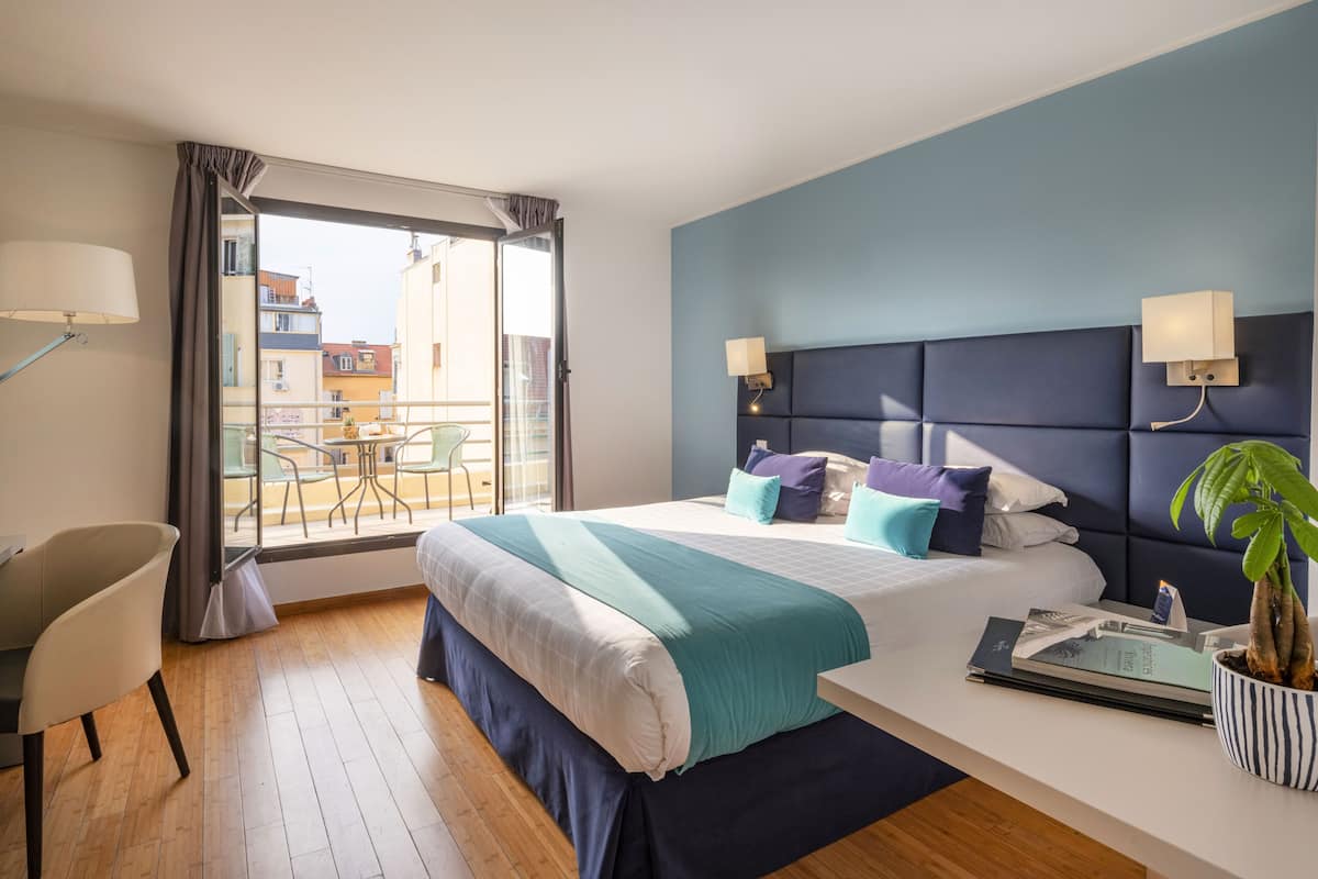 A modern hotel room with a large bed, a desk, and a balcony with chairs.