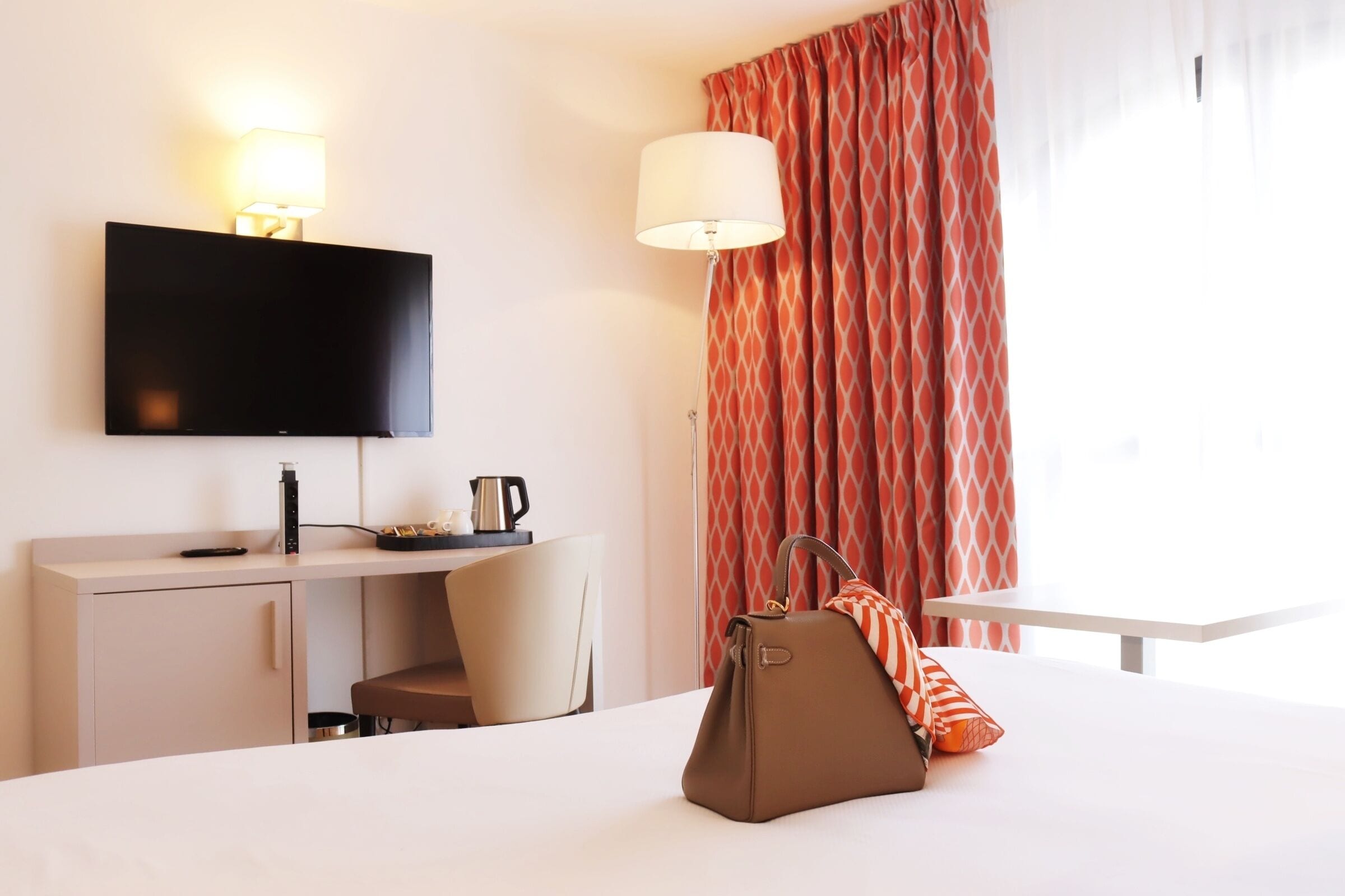 A hotel room with a bed, a desk with a flat-screen TV, a chair, a lamp, and a window with curtains.
