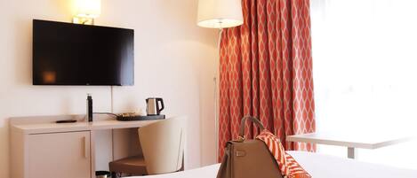 A hotel room with a bed, a desk with a flat-screen TV, a chair, a lamp, and a window with curtains.