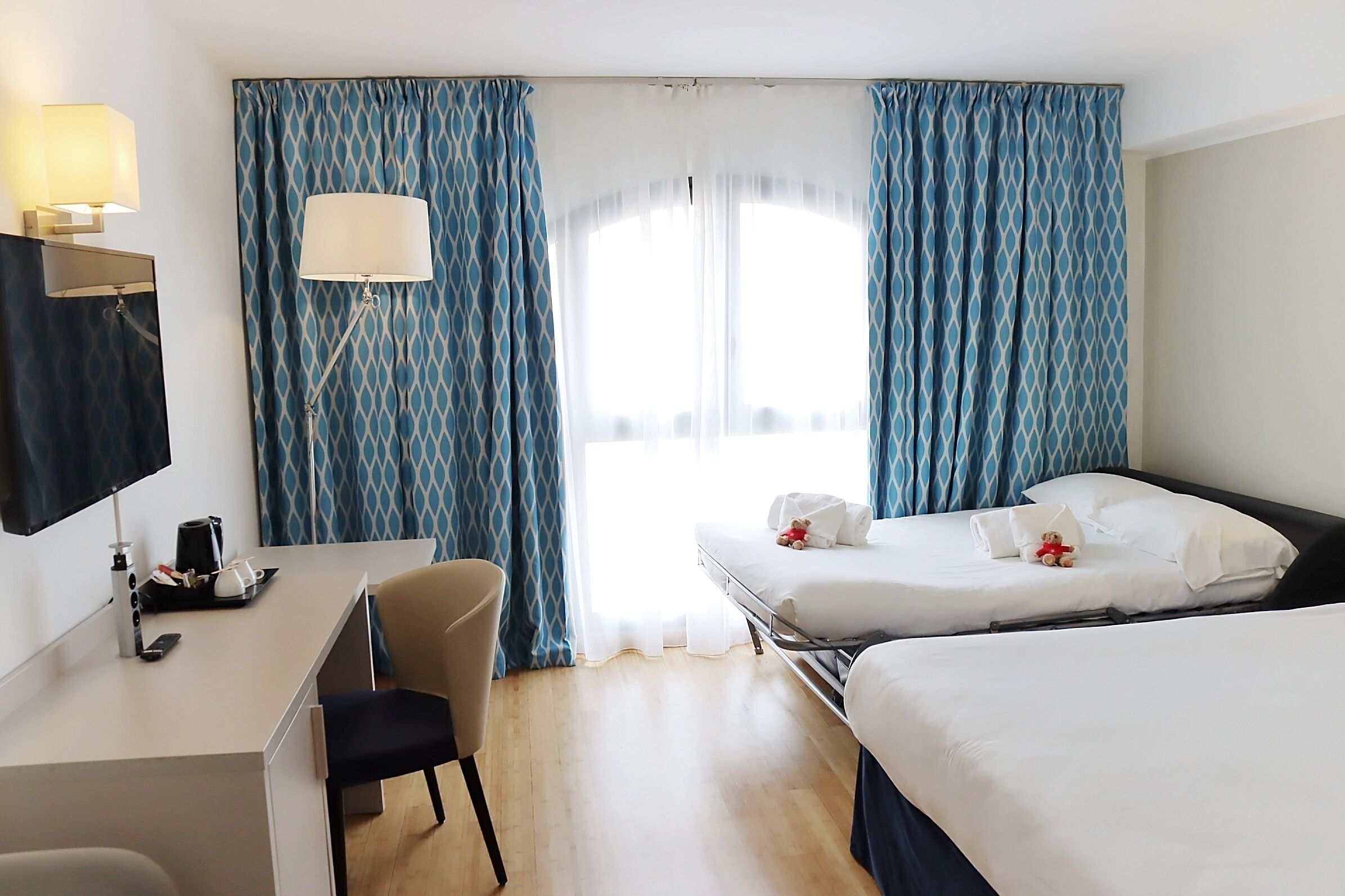 A hotel room with a desk, two beds, a chair, a lamp, and a window with curtains.