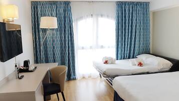 A hotel room with a desk, two beds, a chair, a lamp, and a window with curtains.