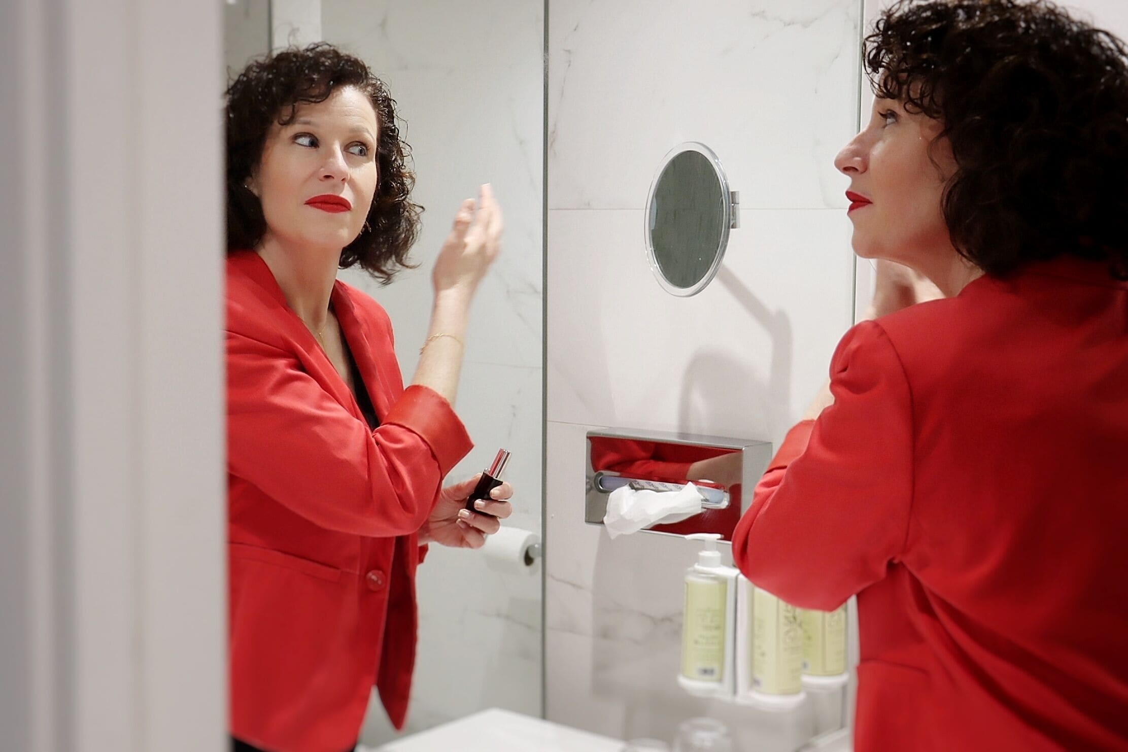 En person i rødt tøj står i et badeværelse med et spejl, en håndklædeholder og toiletartikler.