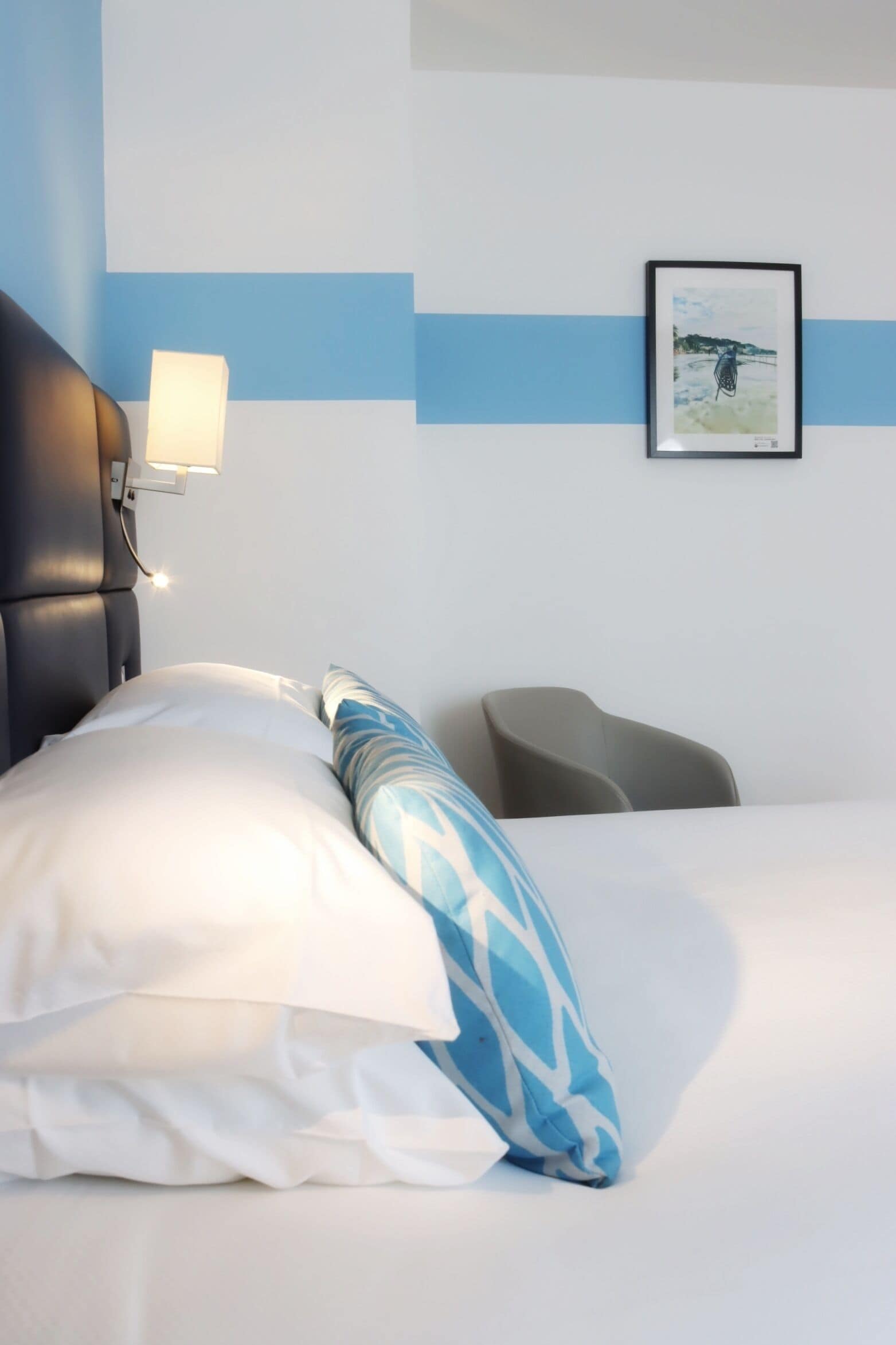 Hotelkamer met een bed, nachtkastje met een lamp, een lijst aan de muur en een stoel.