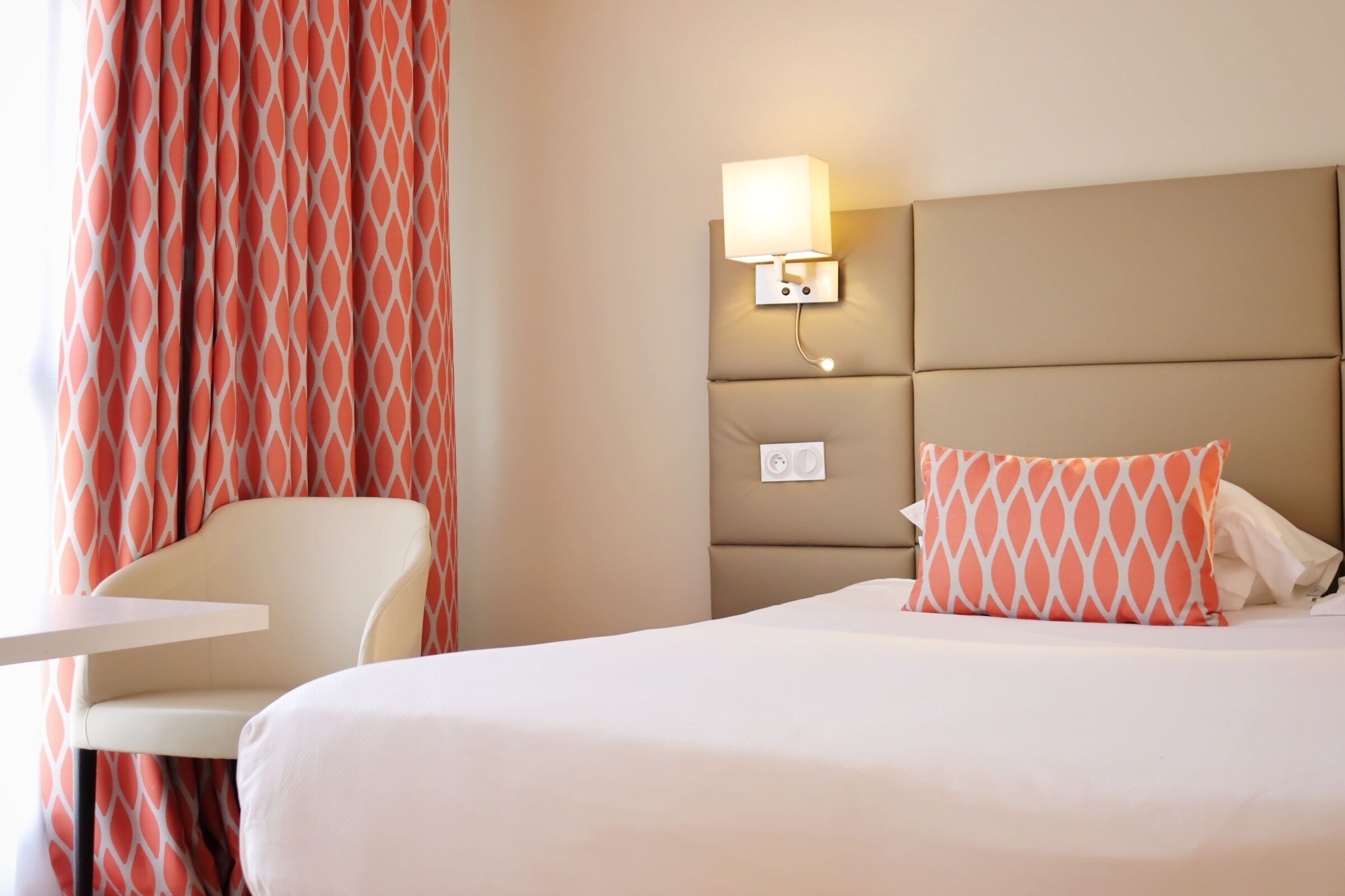 Una habitación de hotel con cama, una silla, una lámpara fijada a la pared y una cortina estampada.