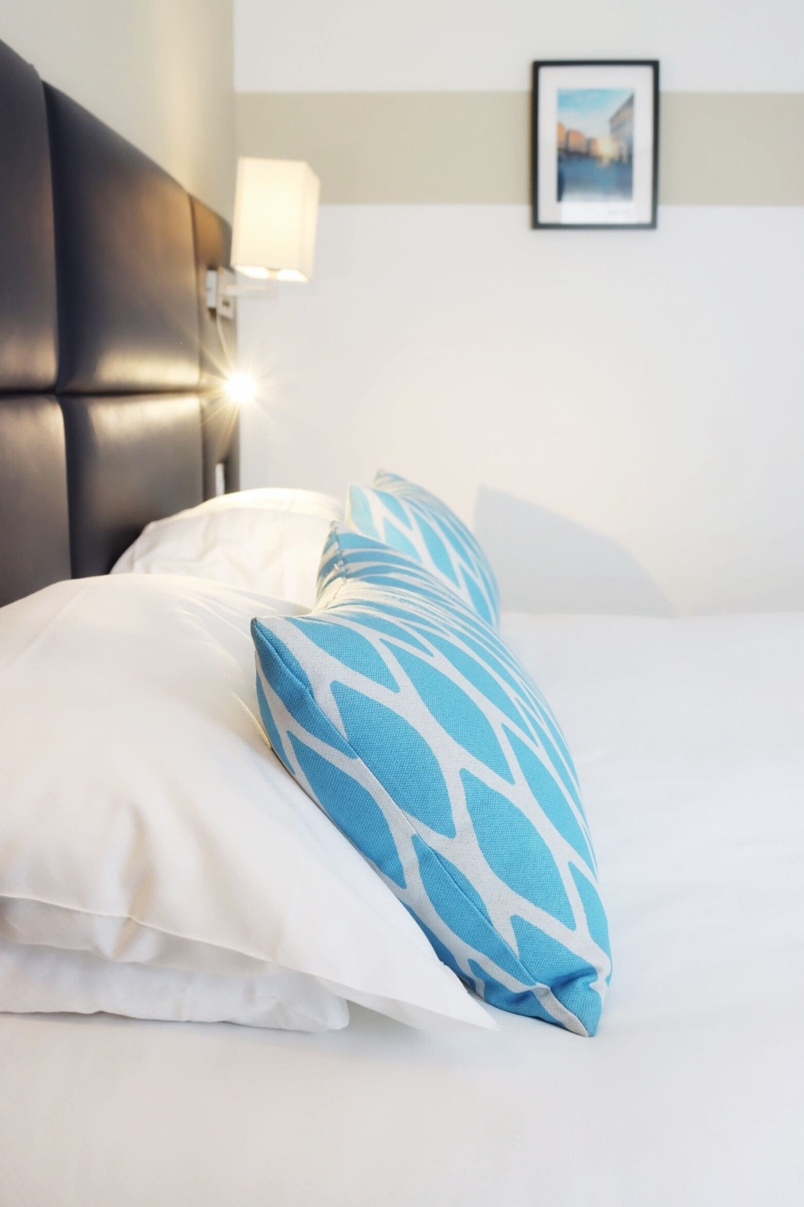 Ein Bett mit Kissen in Weiß und Blau sowie ein schwarzer Kopfteil und eine Wandlampe.