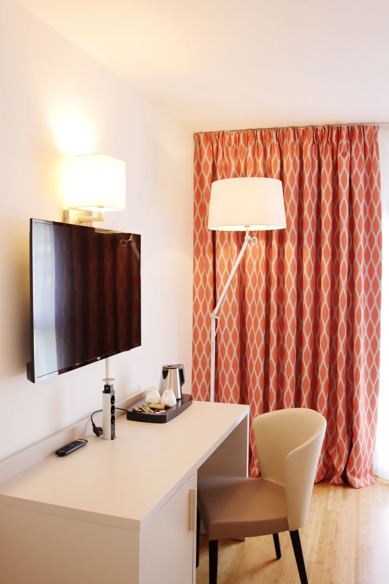 Habitación de hotel con televisión de pantalla plana montada en la pared, un escritorio con lámpara, una silla y una bandeja con tetera y tazas.