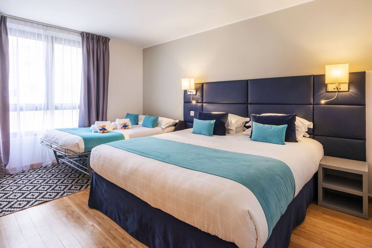 A hotel room with two beds, a large window with curtains, a headboard with built-in lighting, and a bedside table.