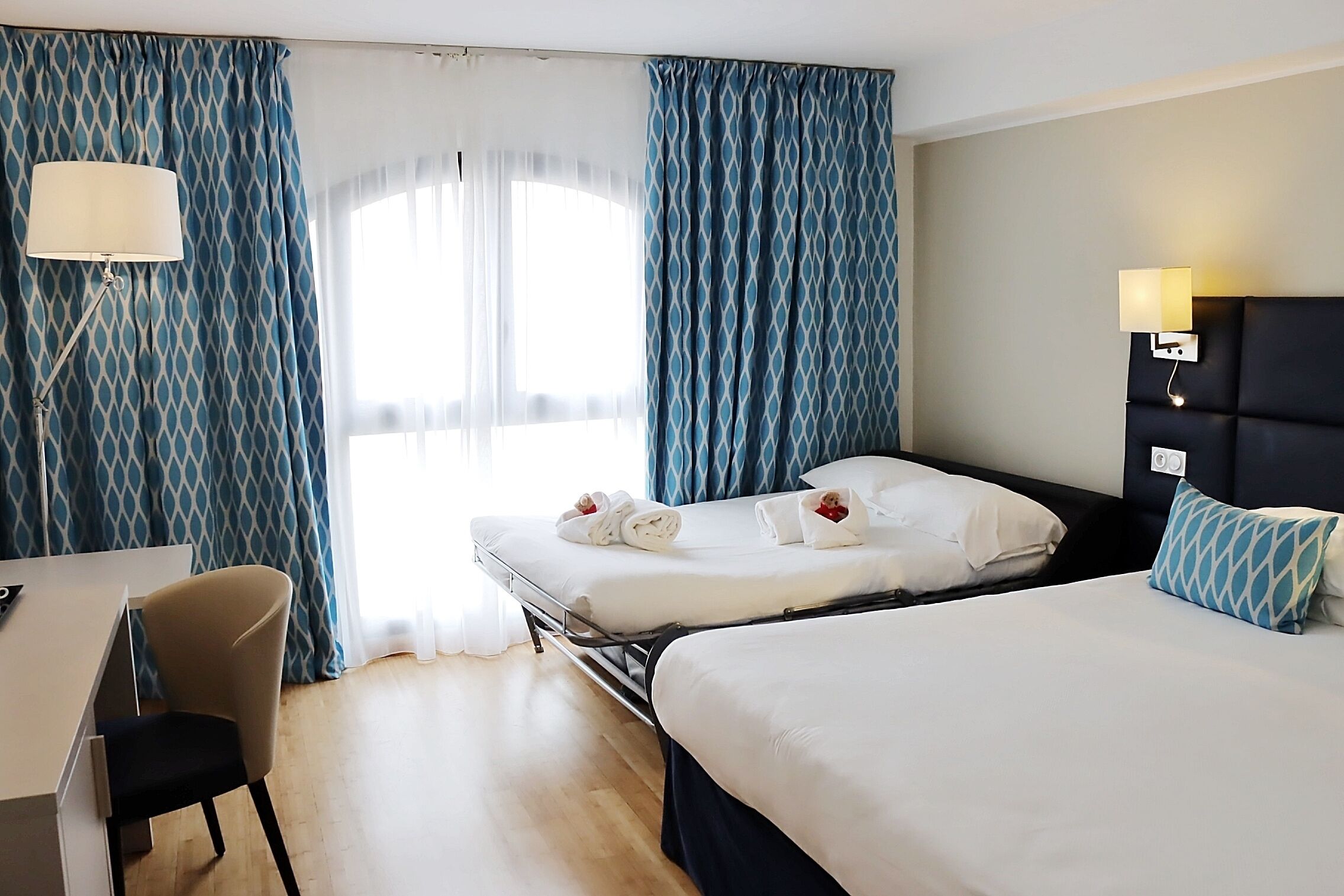 A hotel room with two beds, a desk, a chair, and a window with curtains.