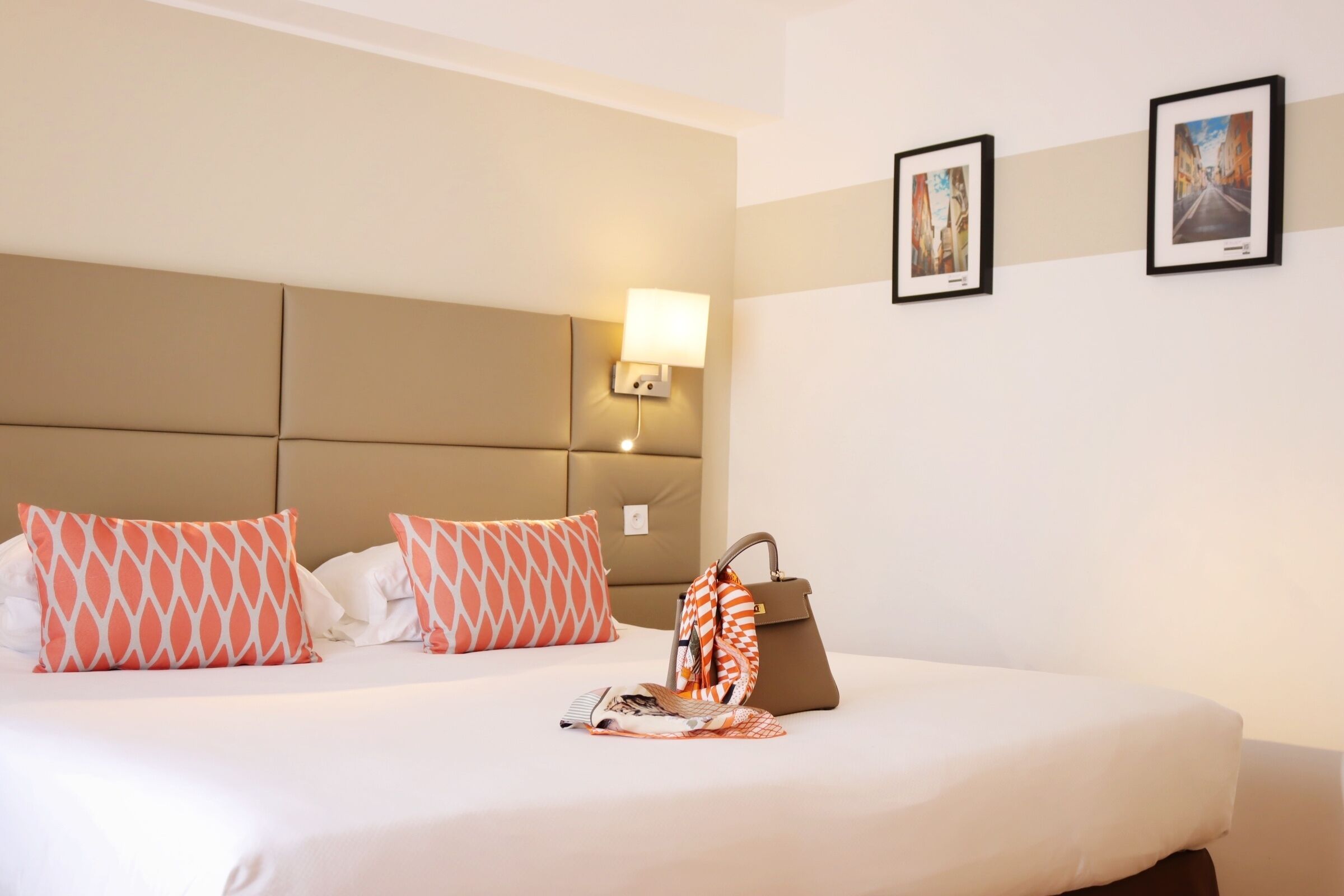 Habitación de hotel con cama, almohadas, mesita de noche, lámpara y dos cuadros en la pared.