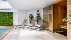 A modern indoor pool area with three sun loungers, a wooden sauna, and a green wall with plants. - Hotel Nice Riviera (Nice)