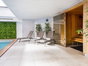 A modern indoor pool area with three sun loungers, a wooden sauna, and a green wall with plants.
