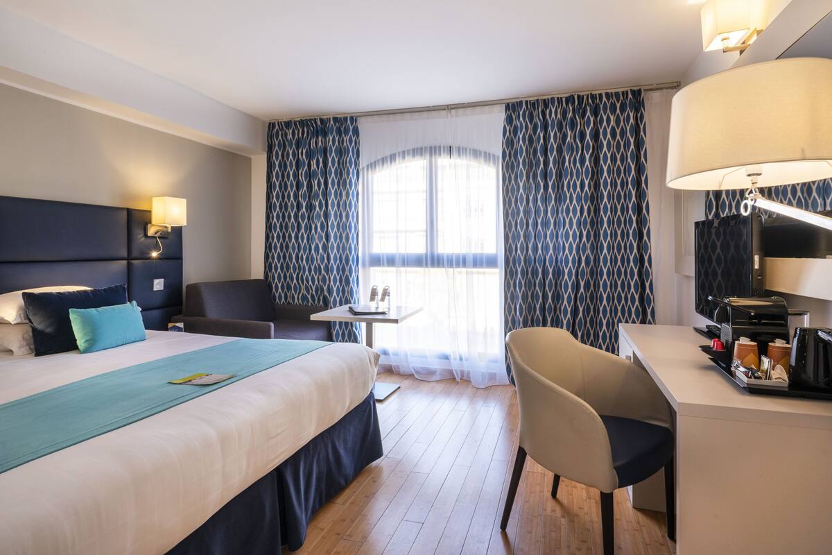 A hotel room with a bed, a desk, a chair, a TV, and a window with curtains.