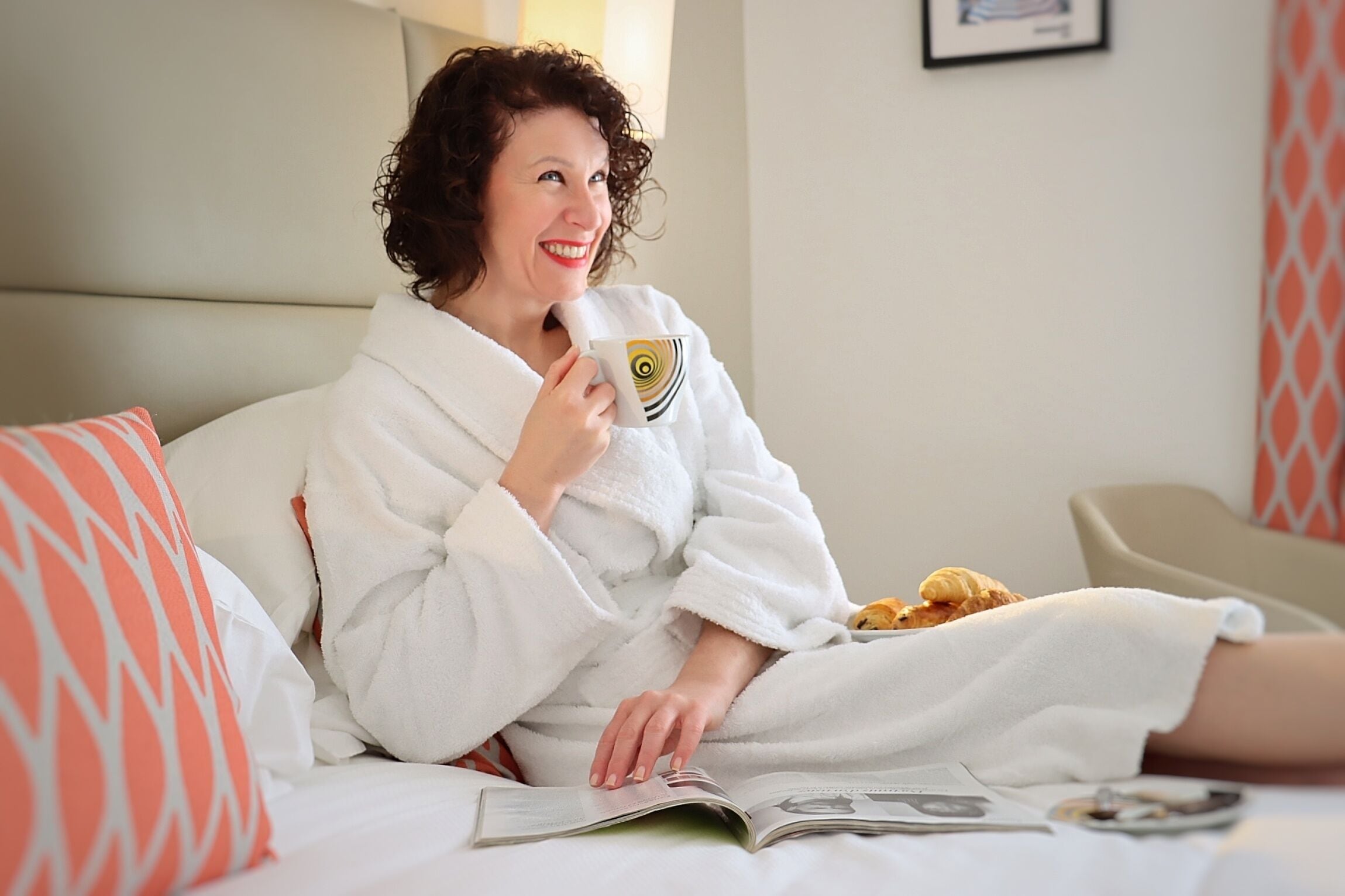 A person in a white bathrobe sitting on a bed with a magazine, a cup, and pastries.