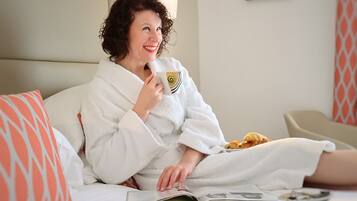 A person in a white bathrobe sitting on a bed with a magazine, a cup, and pastries.