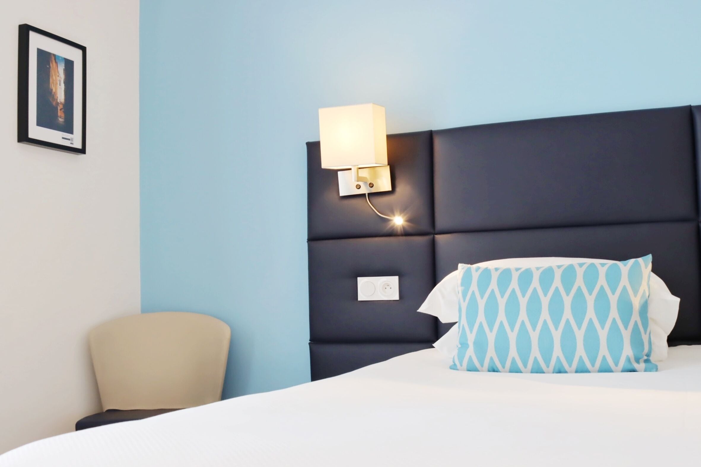 A hotel room with a bed, a nightstand with a lamp, a chair, and a wall-mounted power outlet.