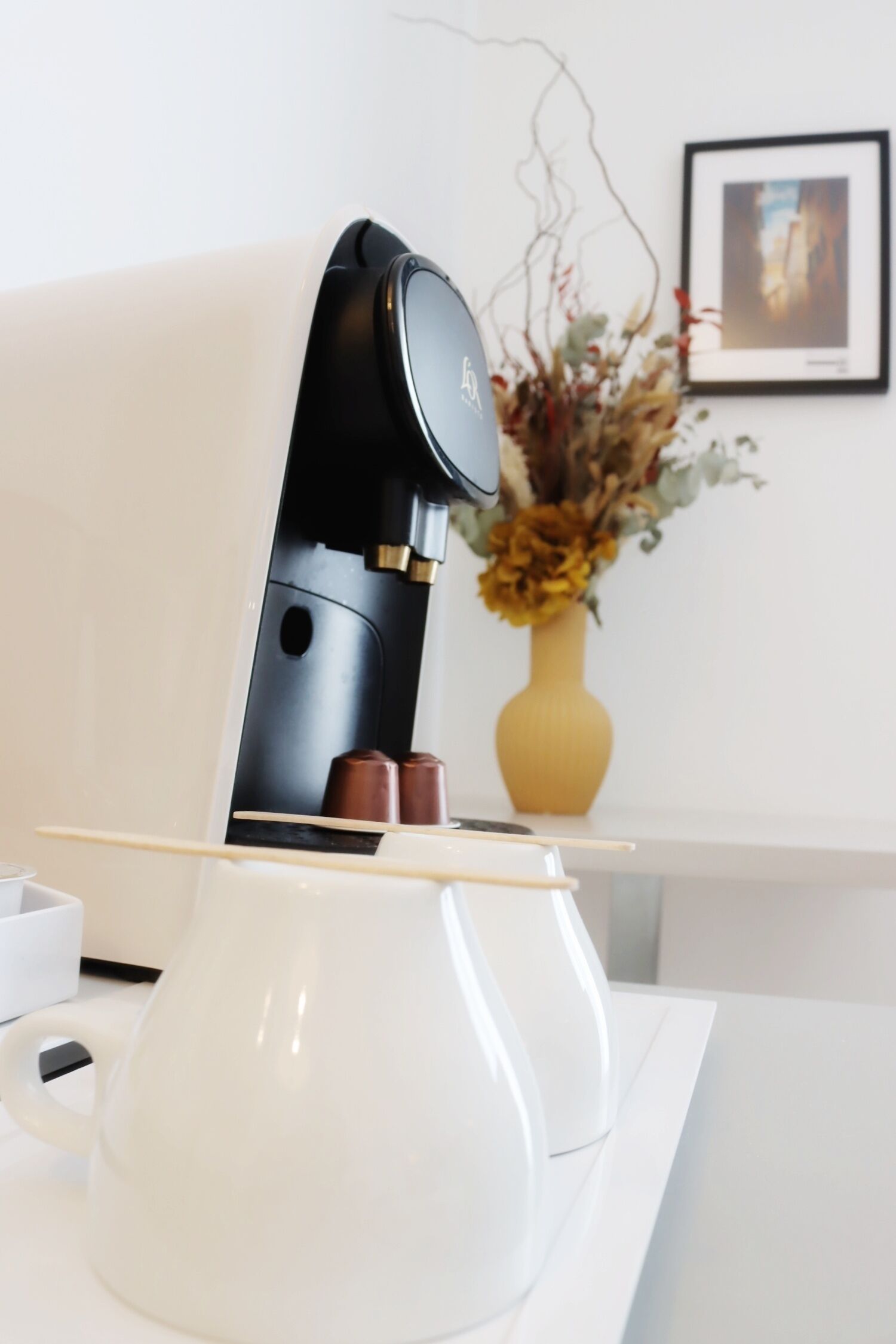 A modern coffee machine with a sleek black design, a white countertop, and a vase with dried flowers in the background.