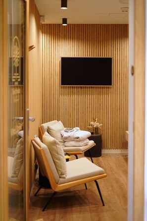 A modern room with a wooden slatted wall, a flat-screen TV mounted above a cushioned chair, and a small side table with a vase.
