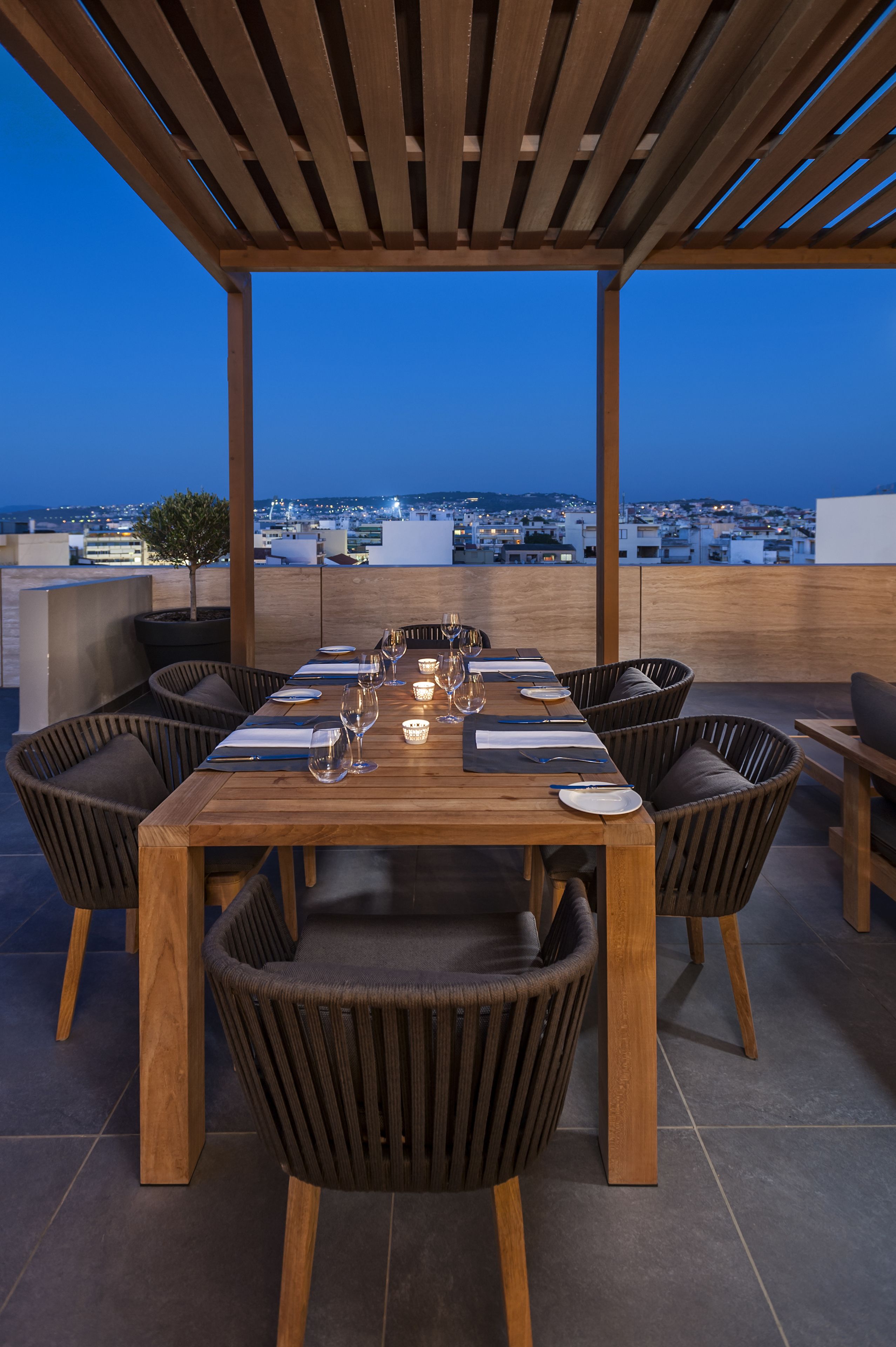Une table de salle à manger en bois, avec six chaises, une plante en pot et une vue sur la ville au crépuscule.