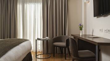 A hotel room with a bed, a desk, two chairs, a small table with a cup and a book, a potted plant, and a TV mounted on the wall.