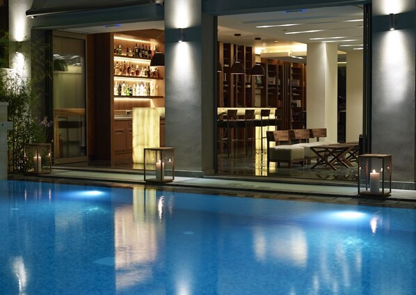 A modern indoor pool area with a bar and seating area, featuring a glass enclosure and ambient lighting. - Samaria Hotel (Chania)