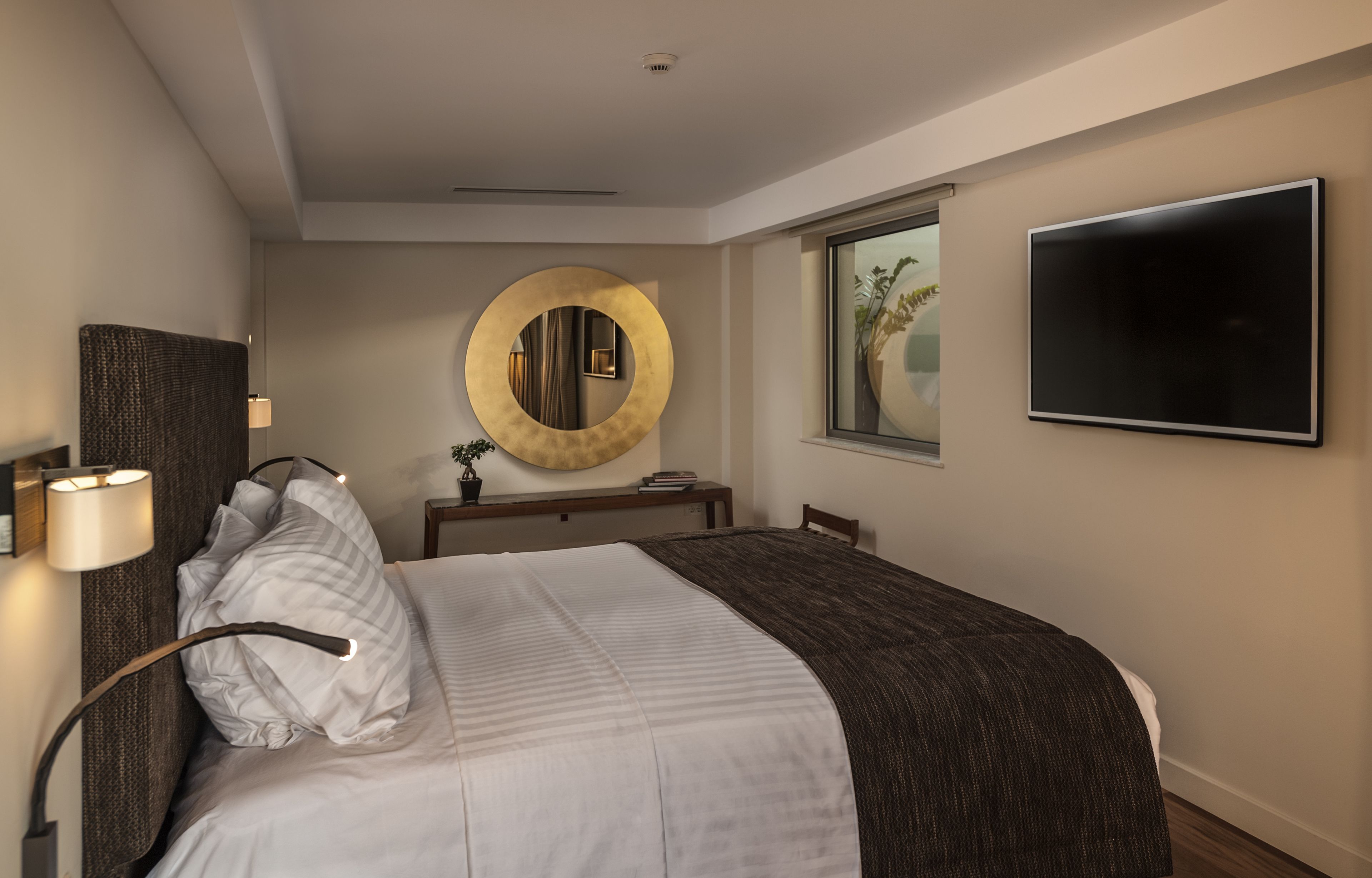 Habitación de hotel con cama, espejo redondo, televisor de pantalla plana, mesita de noche y ventana con vistas a palmeras.