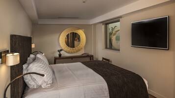 Habitación de hotel con cama, espejo redondo, televisor de pantalla plana, mesita de noche y ventana con vistas a palmeras.
