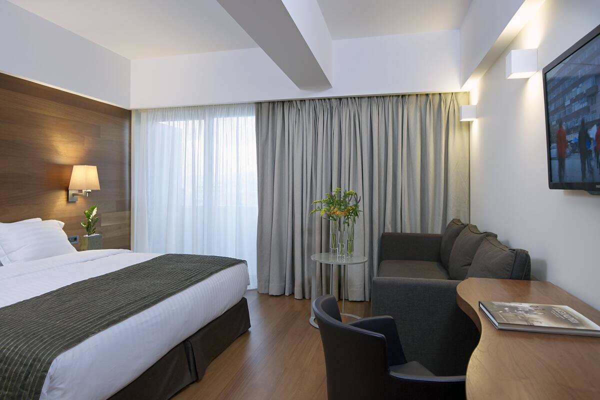 Une chambre d’hôtel avec un lit, un bureau, une chaise, une petite table avec un vase et une grande fenêtre avec des rideaux.