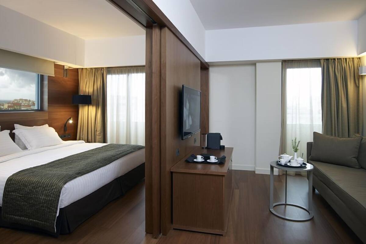 Une chambre d’hôtel avec un lit, une télévision, une petite table avec une tasse et une soucoupe, un canapé et une fenêtre avec des rideaux.