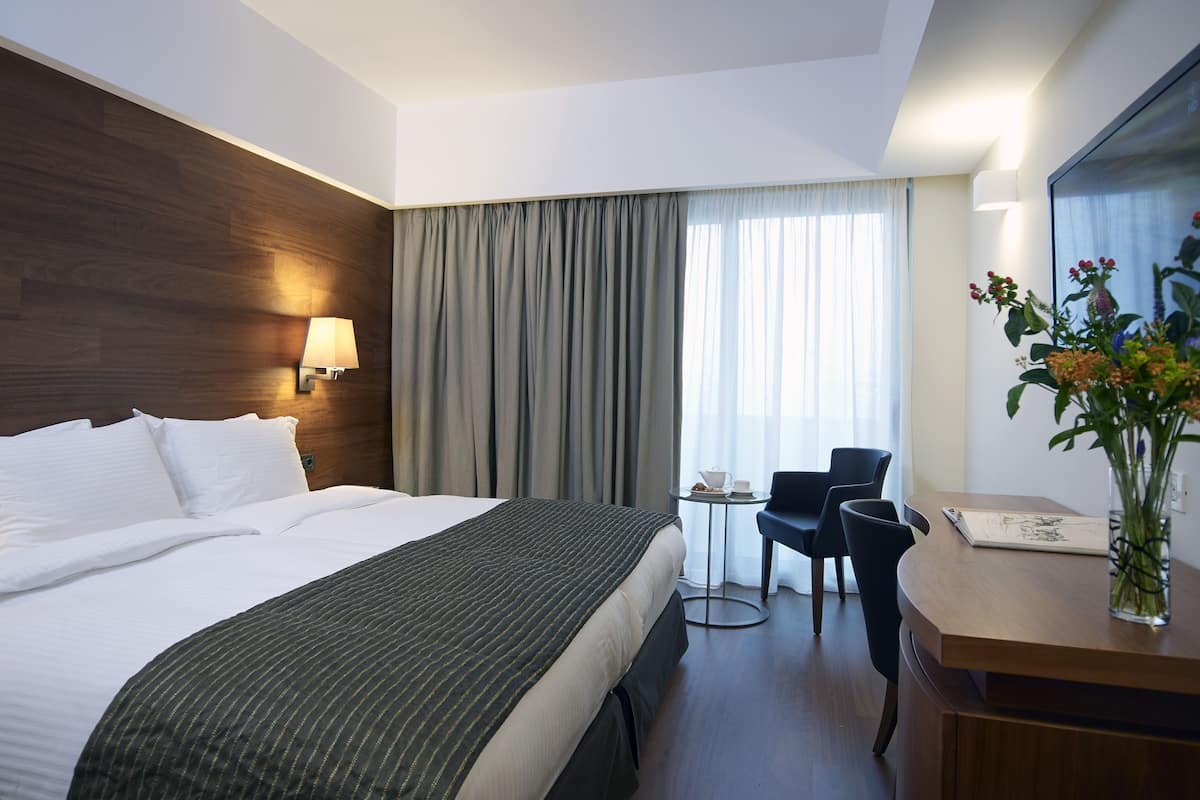 Une chambre d’hôtel avec un grand lit, un bureau avec une chaise, une petite table avec une tasse et une soucoupe, et un vase rempli de fleurs.