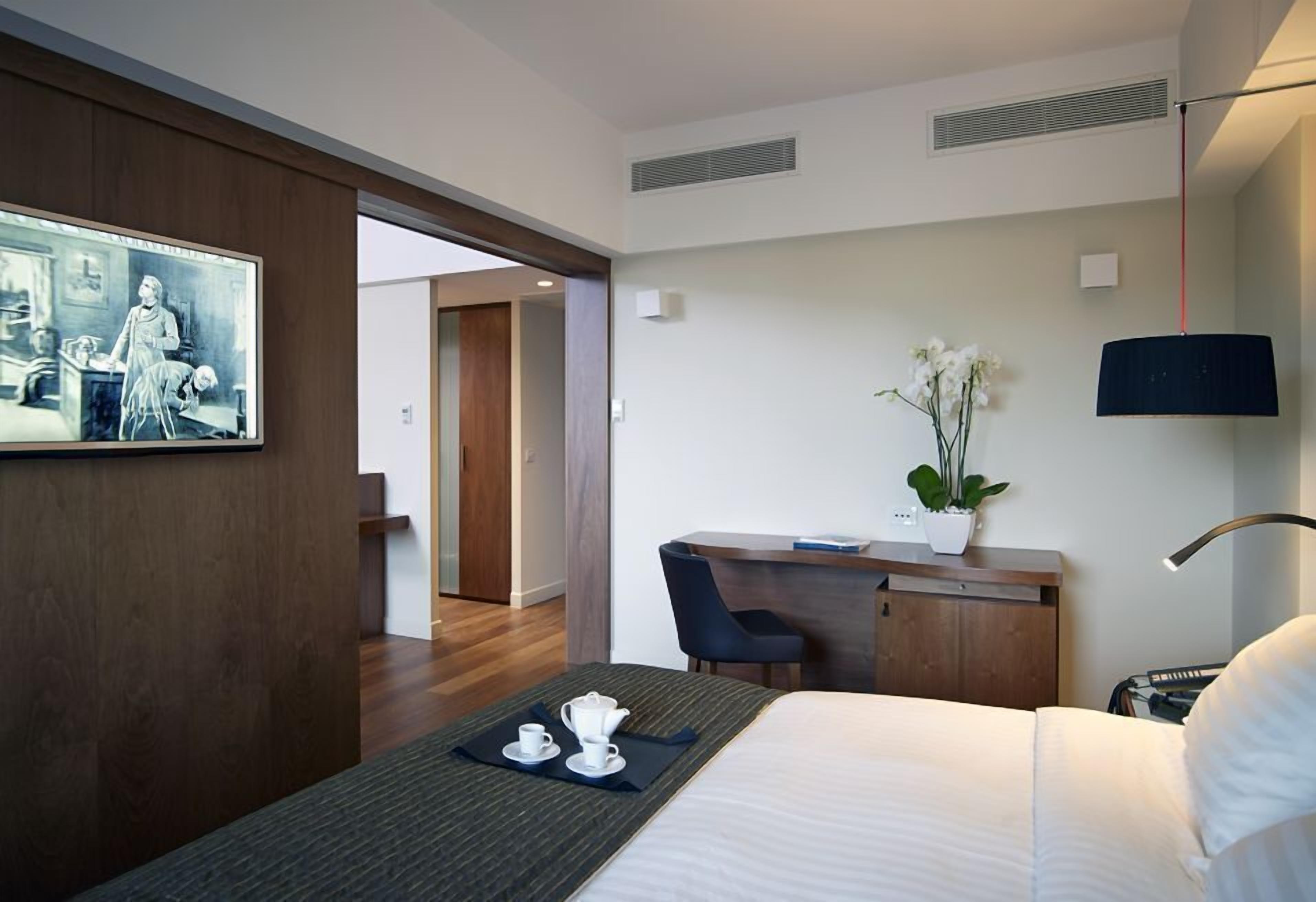 Habitación de hotel con una cama, un escritorio con una silla, una mesita con tetera y tazas, una planta en maceta y un cuadro en la pared.