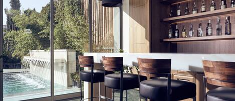 A modern bar area with a glass partition, wooden chairs, and a shelf with bottles.