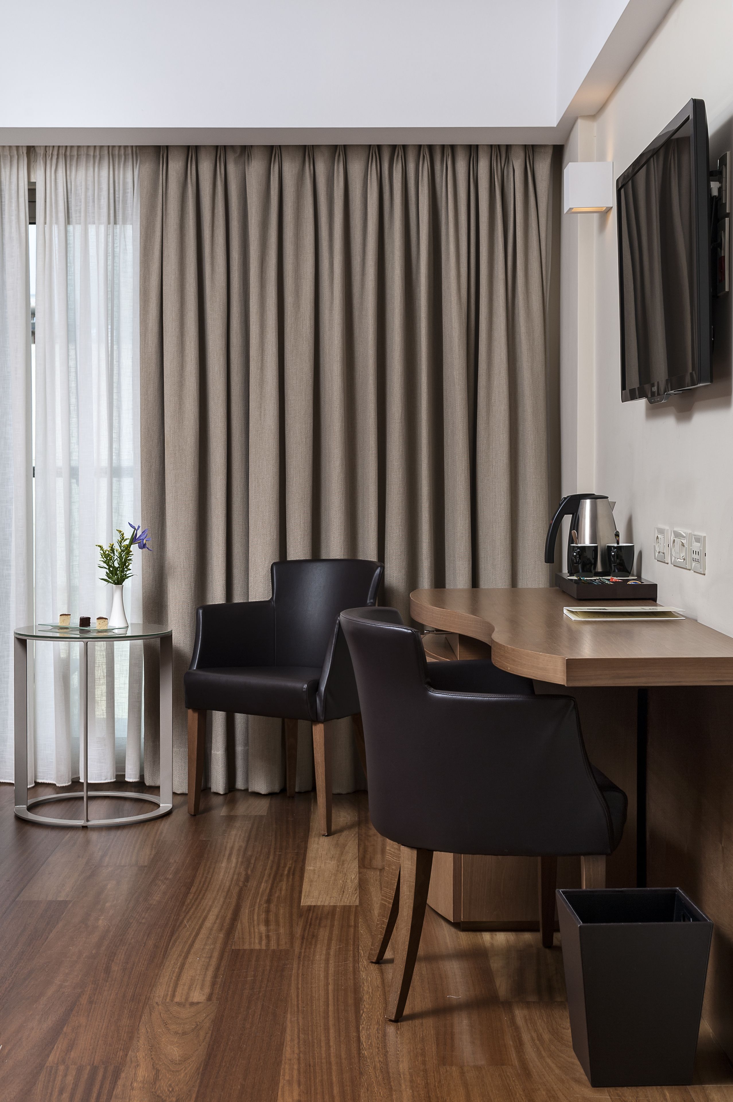 A modern hotel room with a wooden desk, two black chairs, a small side table with a vase, a coffee maker, and a trash bin.
