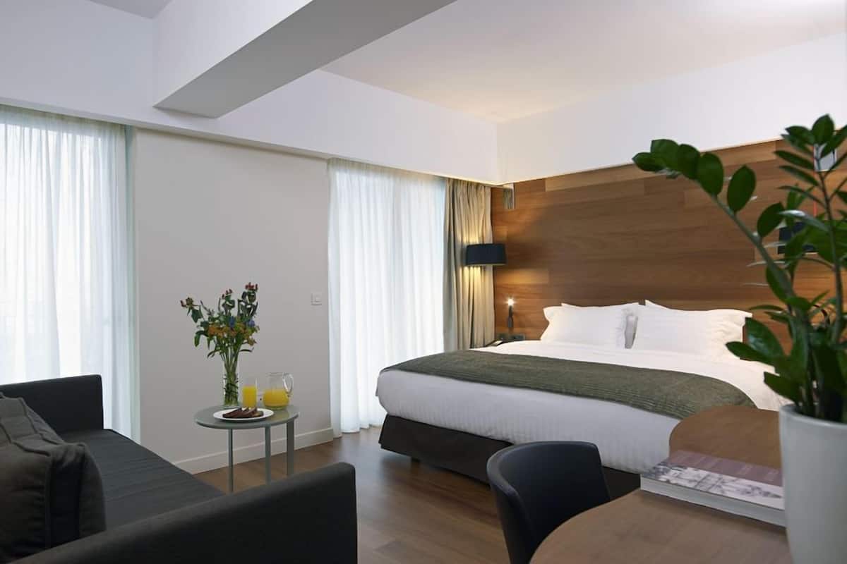 Une chambre d’hôtel moderne avec un grand lit, une petite table ornée d’un vase de fleurs et d’un verre de jus d’orange, un canapé et une tête de lit en bois.