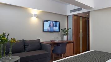 A modern hotel room with a grey sofa, a small desk, a television mounted on the wall, and a potted plant.