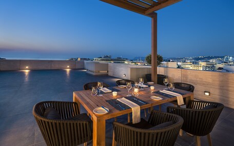 An outdoor dining area with a wooden table set for four, surrounded by wicker chairs, overlooking a cityscape at dusk.