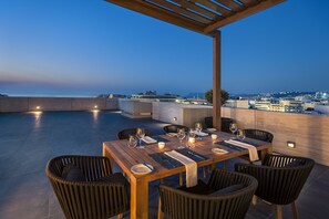 An outdoor dining area with a wooden table set for four, surrounded by wicker chairs, overlooking a cityscape at dusk. - Samaria Hotel (Chania)