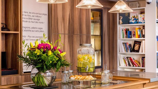 A reception area with a wooden reception desk, a vase of flowers, a glass jar with a drink, and a bookshelf with books and a framed picture. - Hôtel Littéraire Arthur Rimbaud, BW Signature Collection (Paris)