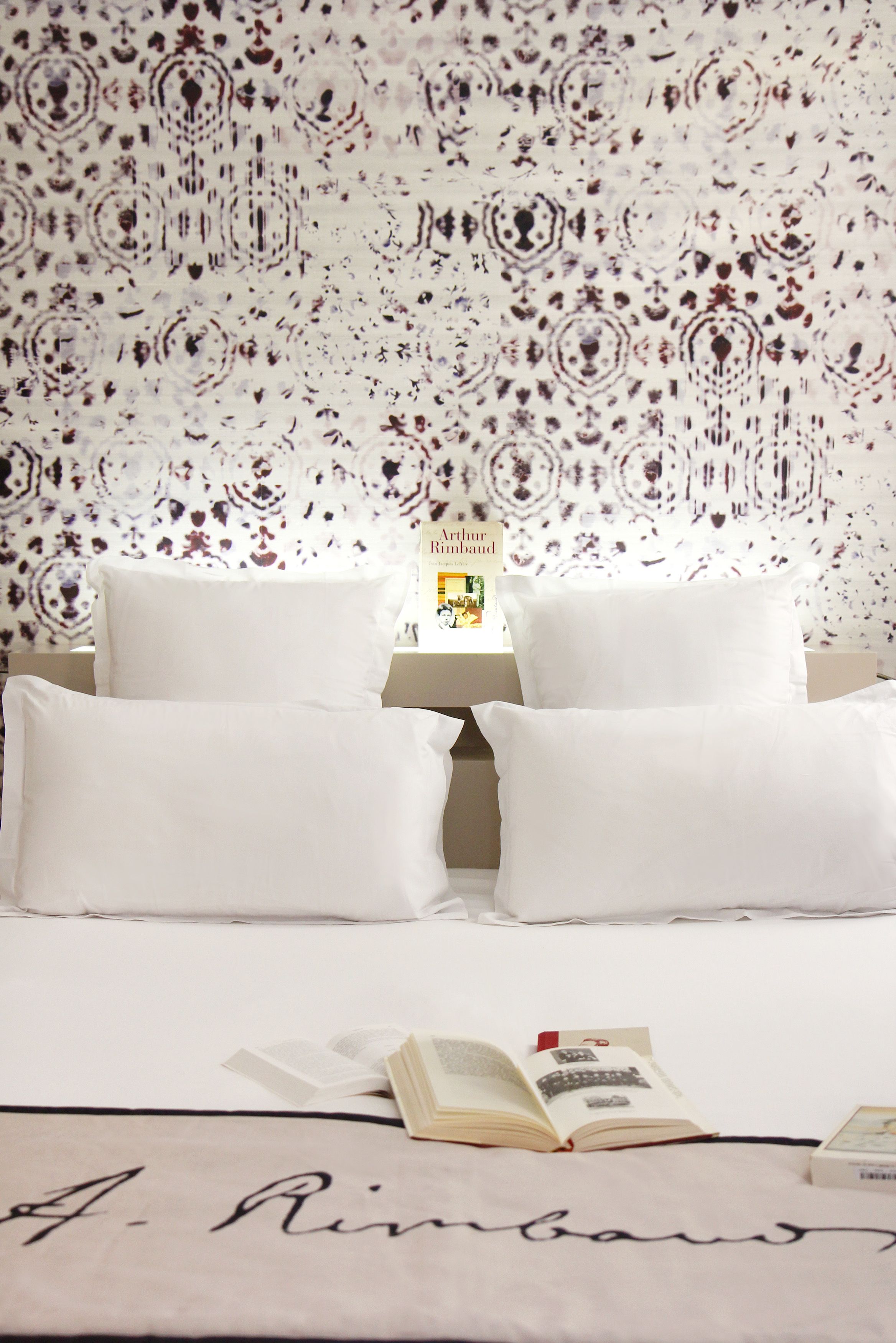 A neatly made bed with white linens and pillows, a book on the bed, and a wall with a patterned wallpaper.