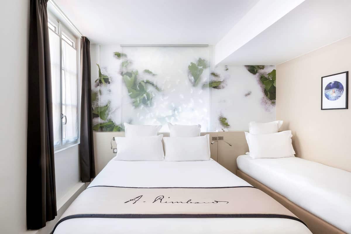 A hotel room with a large bed, a smaller sofa, a window with curtains, and a wall with a plant and a framed picture.