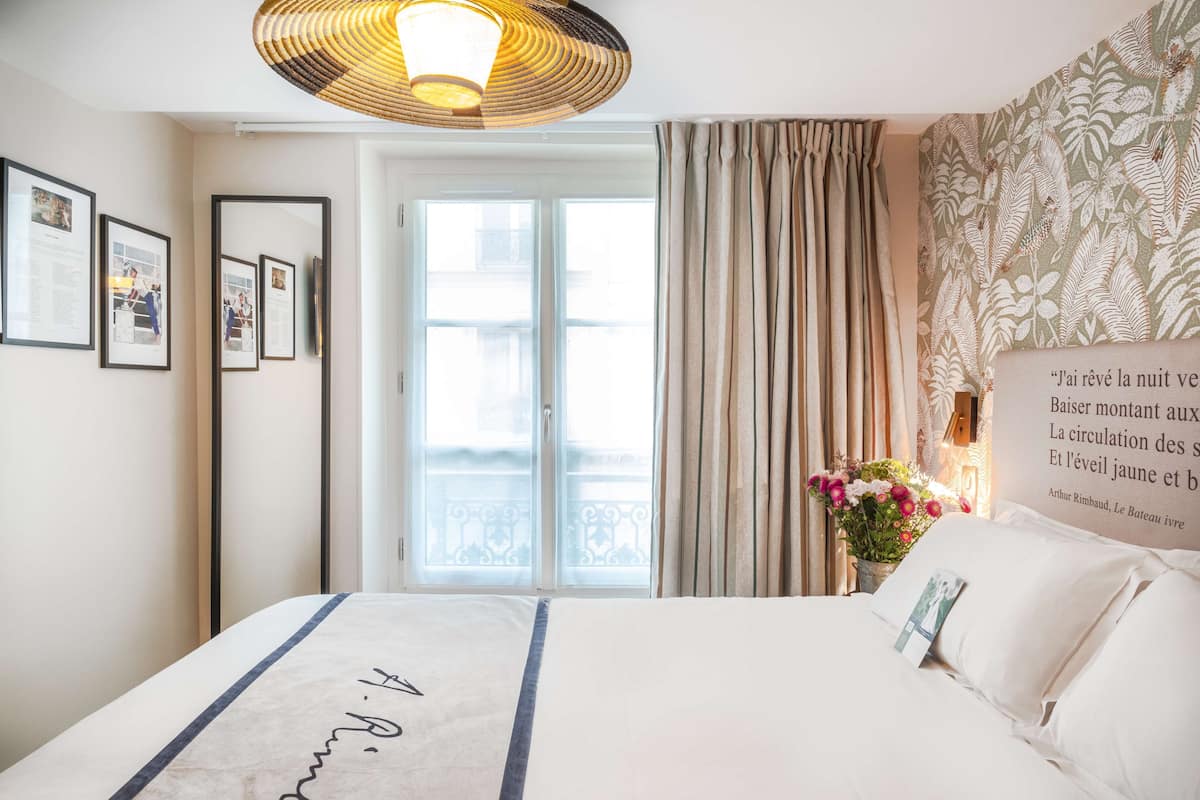A hotel room with a bed, a large window with curtains, a wall-mounted light fixture, and framed pictures on the wall.