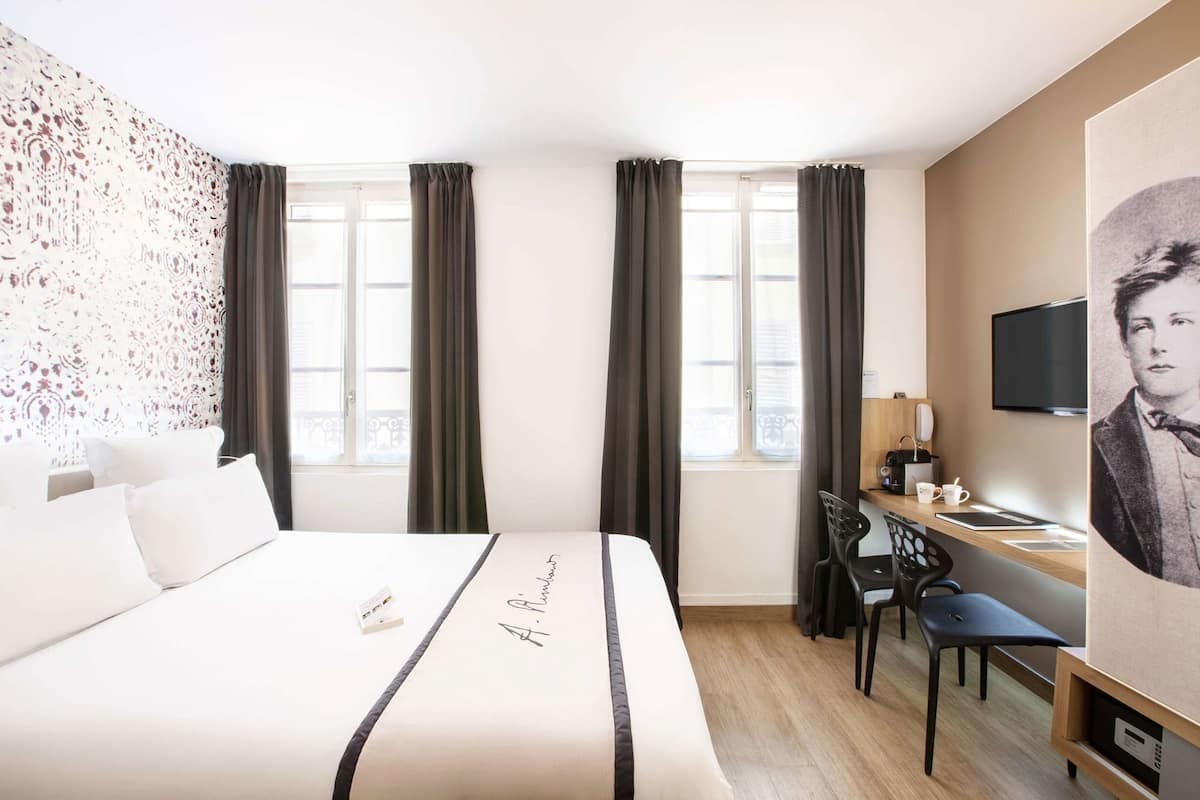 A hotel room with a bed, a desk with a chair, a television, and a window with curtains.
